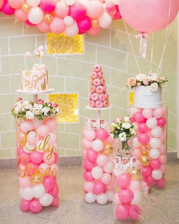 round acrylic plinths with pink and white balloons inside the plinths. Cake, donuts and flowers placed on top of round plinths